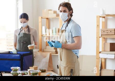 Portrait d'un jeune homme dans un masque de protection utilisant un Tablet pc au travail tout en travaillant dans le service de livraison avec son collègue en arrière-plan Banque D'Images