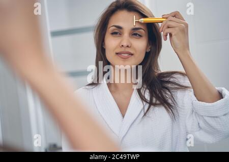 Jeune femme charmante utilisant un masseur pour le visage doré Banque D'Images