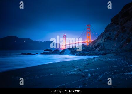 Il vaut la peine de descendre la colline jusqu'à la plage de Bakers et de jeter un coup d'œil au Golden Gate Bridge à l'heure bleue. Le pont rouge semble briller devant le fond bleu. Banque D'Images