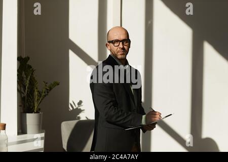 Portrait d'un homme d'affaires mature en lunettes et en costume regardant l'appareil photo tout en remplissant des documents au bureau Banque D'Images