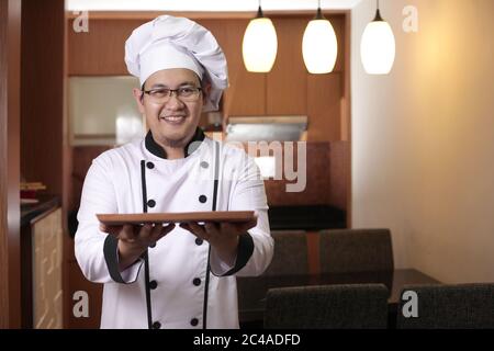 Portrait du chef asiatique masculin semble heureux et fier de présenter quelque chose sur son assiette en bois vide, copier espace repas concept Banque D'Images