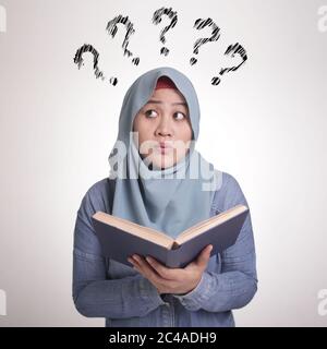 Portrait d'une jeune femme musulmane asiatique portant le hijab, tenant un livre avec expression de pensée et point d'interrogation Banque D'Images