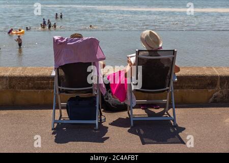 Southend-on-Sea, Royaume-Uni. 25 juin 2020. Deux personnes assises sur des chaises pliantes profitent d'un moment paisible face à la mer depuis la promenade pavée de Westcliff Beach, Essex. Une serviette et un chapeau de soleil suggèrent un temps chaud, tandis que les nageurs et les échassiers en arrière-plan évoquent une atmosphère balnéaire détendue typique de la vie côtière de Southend. Les mesures de distanciation sociale sont assouplies avec des températures qui devraient être supérieures à vingt-cinq cents Penelope Barritt/Alamy Live News Banque D'Images