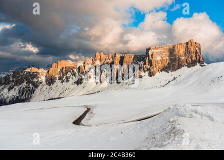 Vue depuis le dessus d'une route sinueuse de montagne à travers les prairies enneigées et sous les hauts sommets rocheux dans les Alpes européennes au coucher du soleil en hiver Banque D'Images