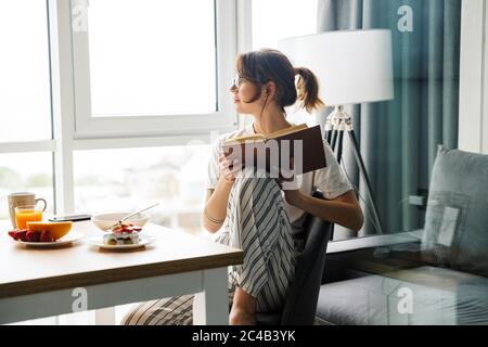 Photo de jeune femme concentrée lisant le livre tout en prenant le petit déjeuner dans une chambre confortable à la maison Banque D'Images