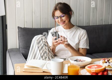 Photo de la femme caucasienne gaie utilisant un téléphone mobile tout en prenant le petit déjeuner dans une chambre confortable à la maison Banque D'Images