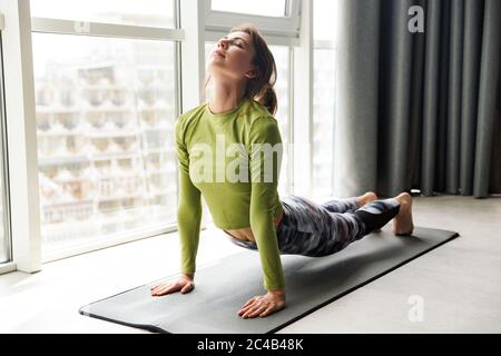 Photo de la femme brunette concentrée faisant des exercices de yoga sur le tapis tout en s'exerçant dans une pièce confortable Banque D'Images