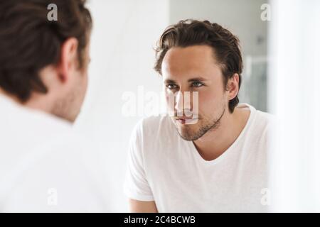 Beau jeune homme confiant debout devant le miroir de salle de bains, regardant lui-même Banque D'Images