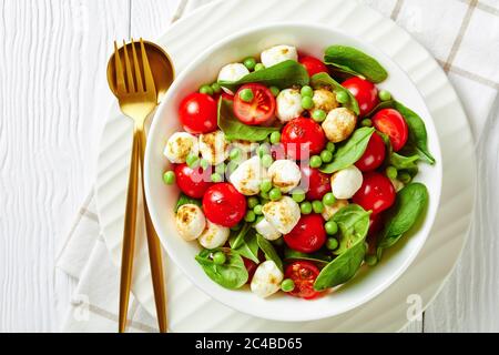 Assiette blanche avec une salade de petits pois frais, mini boules de mozzarella, tomates cerises fraîches aux petits épinards, sauce au vinaigre balsamique sur bois blanc Banque D'Images