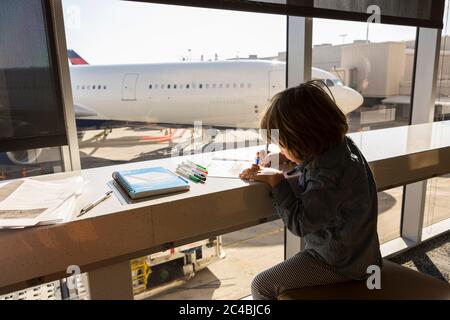 Un jeune de cinq ans qui dessine, assis dans le salon de l'aéroport près d'une fenêtre. Banque D'Images