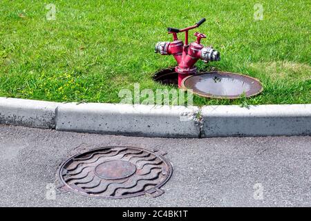 tuyau rouge rouge incendie avec une porte reliée à une trappe ouverte sur une pelouse verte par une route asphaltée avec un trottoir en pierre, par une journée d'été ensoleillée. Banque D'Images