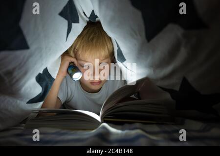 Un petit garçon lit un livre avec une lampe de poche sous les couvertures la nuit. Banque D'Images