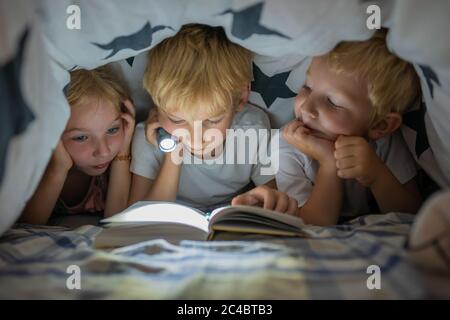 Trois jeunes enfants lisent un livre avec une lampe de poche sous les couvertures la nuit. Banque D'Images