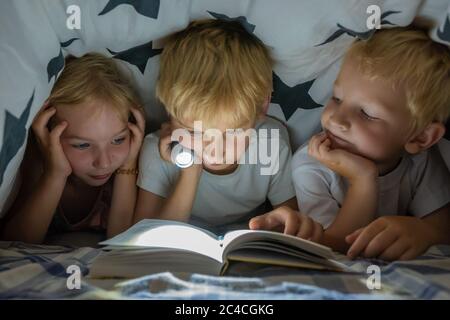 Trois jeunes enfants lisent un livre avec une lampe de poche sous les couvertures la nuit. Banque D'Images