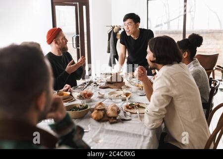 Jeune asiatique en lunettes et T-shirt noir, penché sur la table, discutant avec bonheur avec ses collègues. Groupe d'amis internationaux attrayants dépenser Banque D'Images