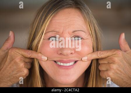 Portrait belle femme mûre avec grand sourire sur l'expression faciale, les doigts s'étirant la bouche au grin, arrière-plan flou. Banque D'Images