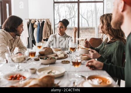 Souriante femme afro-américaine aux cheveux fclés sombres, assise à la table, discutant avec ses collègues de façon rêveuse. Groupe de jeunes amis internationaux pendant Banque D'Images