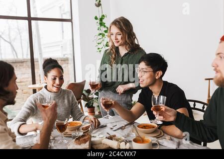 Belle femme aux cheveux ondulés tenant un verre de vin à la main debout près de la table pleine de nourriture. Groupe d'amis internationaux attrayants heureusement Banque D'Images