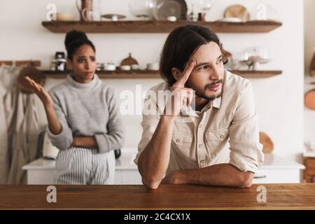 Homme en colère avec une barbe penchée sur la table, tout en regardant malheureusement de côté avec une femme afro-américaine émotive sur le fond. Jeune couple international Banque D'Images