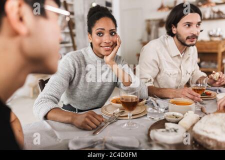 Femme afro-américaine souriante aux cheveux fclés foncés assise à la table penchée sur la main et regardant l'homme. Groupe de jeunes internationaux Banque D'Images