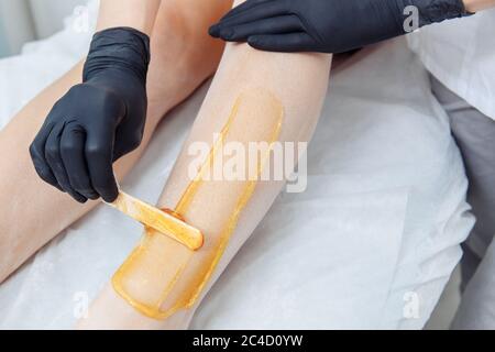 Une main dans un gant en latex noir applique de la cire jaune sur une jambe non rasée. Banque D'Images