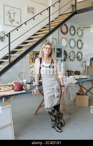 Portrait d'un artiste mature et attrayant avec des cheveux blonds portant des lunettes et un tablier debout dans un studio moderne Banque D'Images