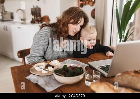 Jeune belle femme avec cheveux rouges en tricot pull et son petit fils assis à la table avec de la nourriture et regardant des dessins animés sur ordinateur portable. Dépenses de maman Banque D'Images