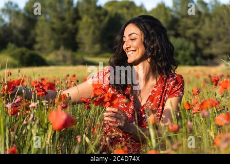 Jeune femme brune vêtue d'une robe rouge avec des imprimés croquant au milieu d'un champ de coquelicots touchant les fleurs tout en souriant Banque D'Images