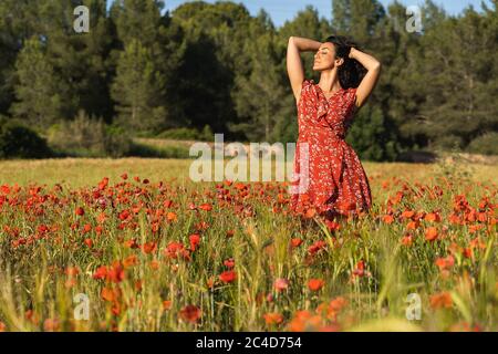 Jeune femme brune vêtue d'une robe rouge avec des empreintes au milieu d'un champ de pavot avec ses mains dans ses cheveux et une expression détendue entourée d'un Banque D'Images