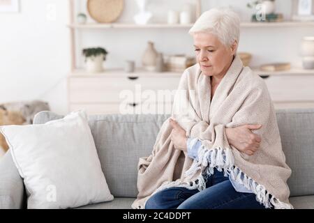 Femme de haut niveau assise sur un canapé recouvert d'une couverture, frississant avec une température élevée Banque D'Images