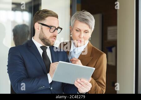 Homme d'affaires en lunettes et en costume montrant quelque chose à une femme d'affaires mûre sur tablette numérique Banque D'Images