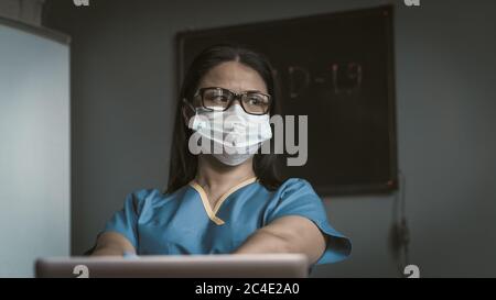 Le Medic fatigué se présente de manière réfléchie à côté. Une femme pensive pense s'asseoir au bureau avec un ordinateur portable. Concept de pandémie. Image en tons Banque D'Images