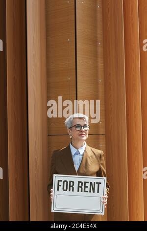 Portrait d'une femme d'affaires mature dans des lunettes tenant l'étiquette de location tout en se tenant contre le bâtiment de bureau Banque D'Images