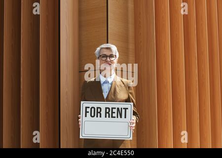 Portrait d'un agent immobilier mature dans des lunettes tenant un écriteau pour louer et sourire à l'extérieur de la caméra Banque D'Images