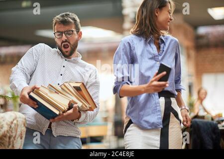 un vieil homme qui porte une pile lourde de livres qui se retombera des mains. une jeune femme qui regarde loin avec son téléphone cellulaire dans ses mains Banque D'Images