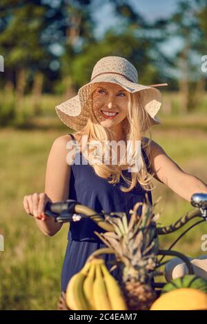 Belle femme blonde avec un sourire sympathique debout à l'extérieur avec son vélo au soleil de printemps portant un chapeau de paille à large bord Banque D'Images