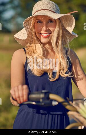 Belle femme blonde avec un sourire sympathique debout à l'extérieur avec son vélo au soleil de printemps portant un chapeau de paille à large bord Banque D'Images