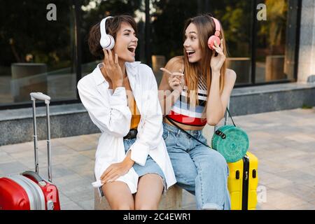 Photo de deux belles femmes heureux d'écouter la musique au casque et de chanter alors qu'il était assis avec des valises au city street Banque D'Images