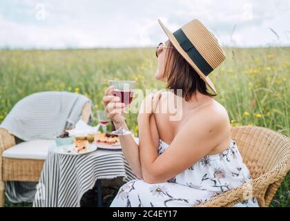 Jeune femme en lunettes de soleil et chapeau de paille habillé léger robe d'été assis dans un fauteuil en rotin sur la prairie haute herbe verte près de table de pique-nique gaie, Banque D'Images