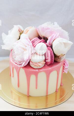 Petit gâteau d'anniversaire avec chocolat rose fondu, fleurs fraîches, macarons et meringues sur fond blanc. Banque D'Images