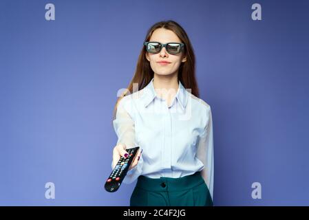 Une femme heureuse dans une chemise bleue regarde des films et des émissions de télévision à la télévision avec des lunettes 3D. Belle fille dans le studio sur un fond lilas commute c Banque D'Images