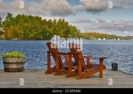 Vie à la retraite - deux chaises de Muskoka assises sur un quai à bateaux face à un lac calme avec arbres et chalets Banque D'Images