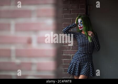 Portrait d'une fille aux cheveux verts en robe acclamée avec maquillage de joker sur un mur de brique flou d'arrière-plan. Banque D'Images