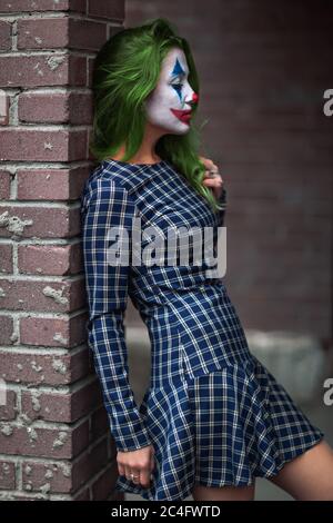 Portrait d'une fille aux cheveux verts en robe acclamée avec maquillage de joker sur un mur de brique flou d'arrière-plan. Banque D'Images