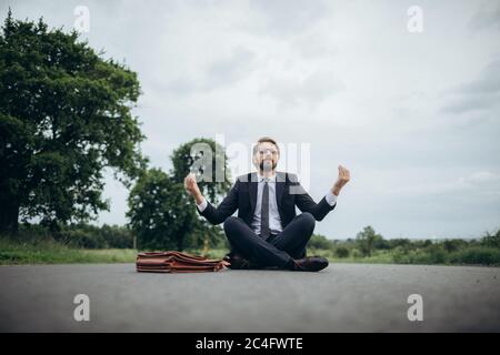 Homme barbu en costume noir assis dans lotus pose sur la route Banque D'Images