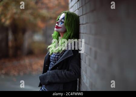 Portrait d'une fille aux cheveux verts en robe acclamée avec maquillage de joker sur un mur de brique flou d'arrière-plan. Banque D'Images