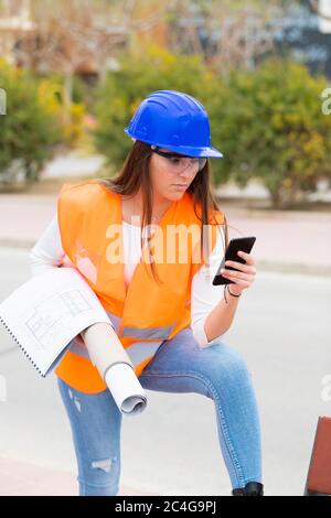 Une jeune femme très belle et concentrée, portant un gilet réfléchissant, un casque de sécurité et des lunettes, vérifie son téléphone en cas de pause. Concept de travail et d'apprentissage. Banque D'Images