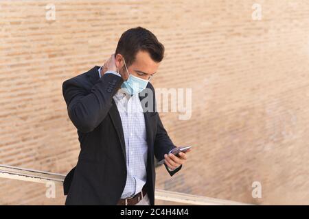 Homme d'affaires avec une veste bleue et un masque sur son visage faisant un geste inquiet tout en regardant son mobile devant un mur de briques Banque D'Images