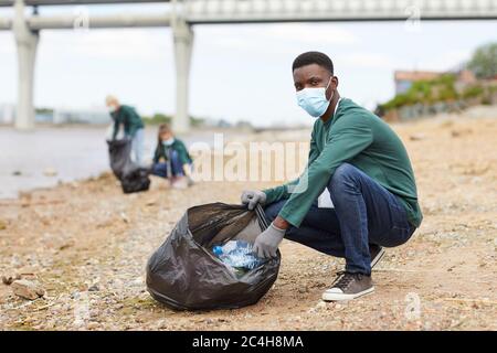 Portrait d'un jeune homme africain en masque de protection regardant l'appareil photo tout en mettant les ordures dans le sac sur la rive de la rivière Banque D'Images