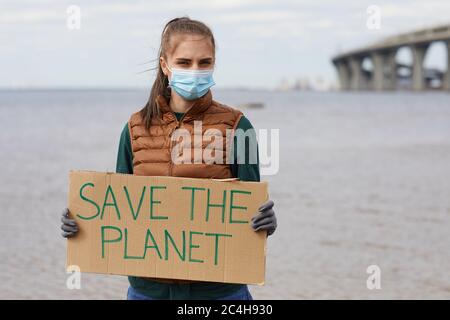 Portrait de la jeune femme dans le masque de protection tenant l'écriteau sauver la planète et regarder l'appareil photo tout en se tenant près de la mer Banque D'Images